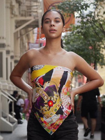 Model wearing the Salute to Youth 90 silk scarf by Cult Luxury, styled as a bandeau top with a vibrant crown and floral collage design, against an urban background.