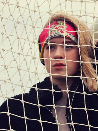 Model styled with the Cult Luxury silk accessory, Butterfly Season hand-rolled silk scarf 65, shot behind a net, highlighting its vibrant red, yellow, and floral patterns